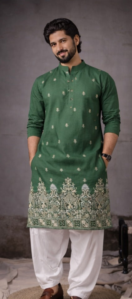 A man modeling a green kurta with white embroidery and white pants, standing against a textured gray studio background.