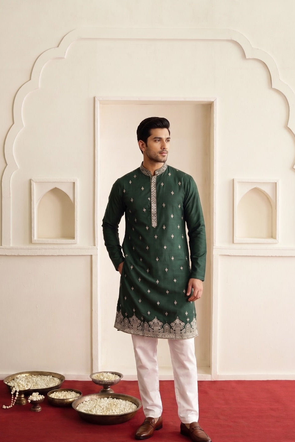 A man modeling a dark green kurta with silver embroidery and white trousers, standing in front of a cream-colored architectural wall with a red carpet. A text overlay 'MKB5104' is visible in the bottom-left corner.