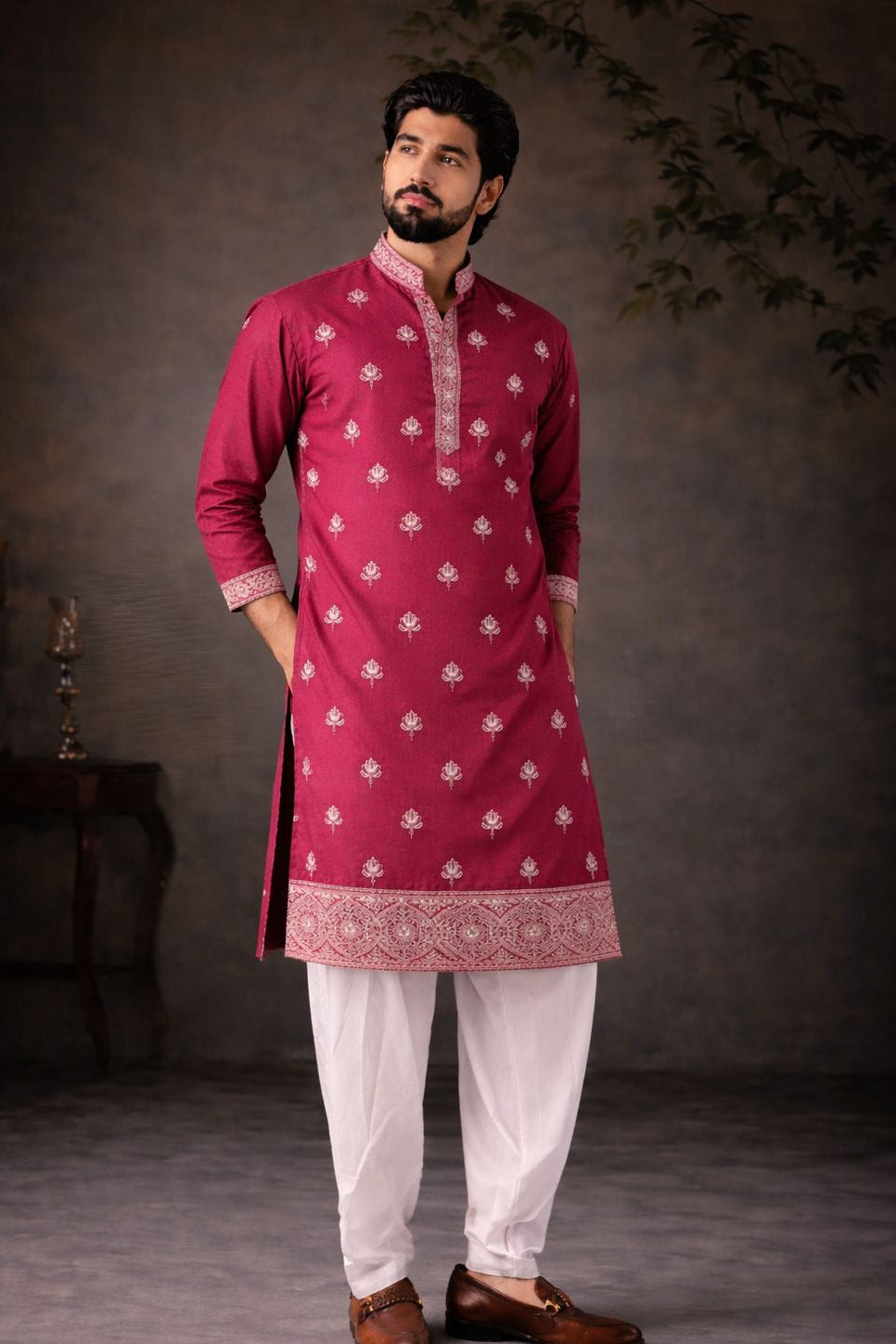 A man modeling a magenta pink kurta with white floral embroidery, paired with white pants and brown loafers, standing against a dark textured studio backdrop.