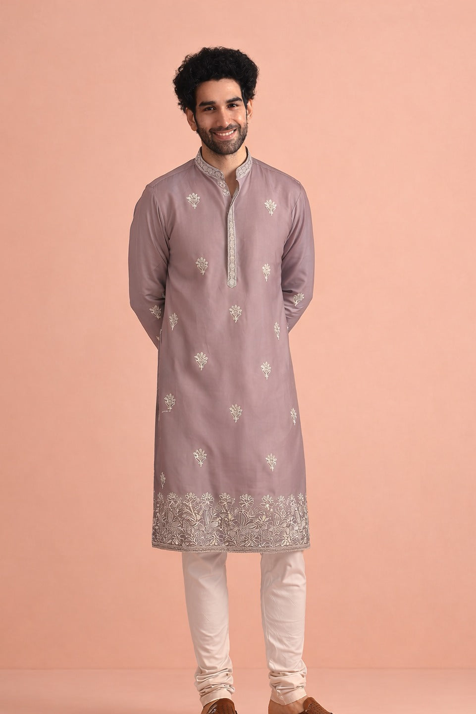 A man modeling a light purple/lavender kurta with white floral embroidery and a silver patterned border, paired with off-white trousers and brown shoes, against a solid peach-pink background.