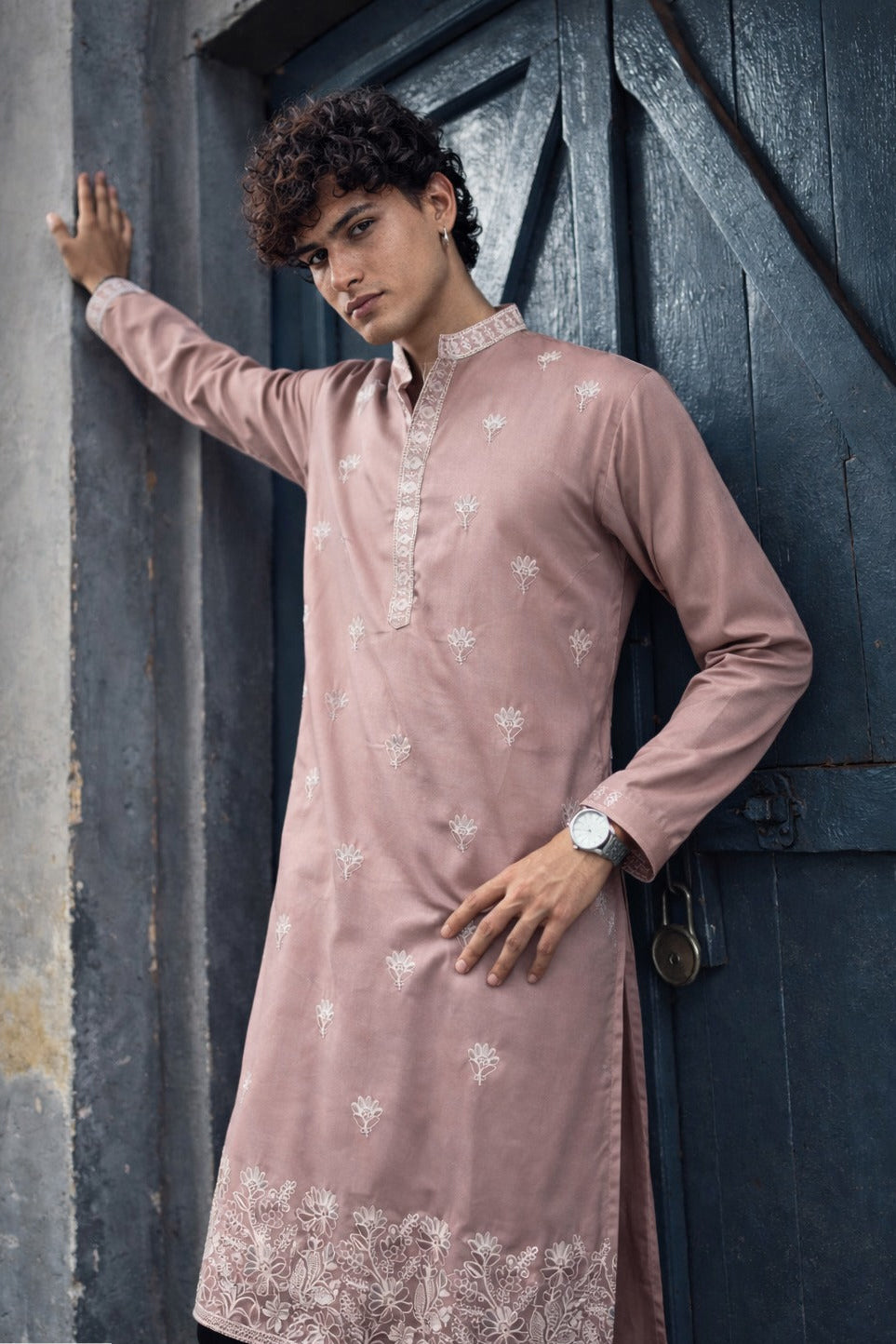 A man wearing a dusty pink long-sleeve kurta with white floral embroidery and a lace hem, posing against a dark wooden door.