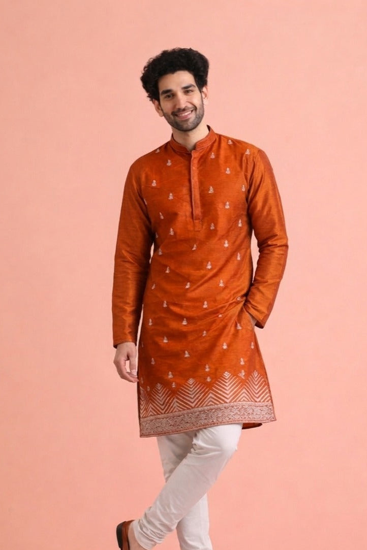 A man modeling a rust-orange kurta with white printed motifs, paired with white trousers and brown leather shoes, standing against a solid peach-pink background.