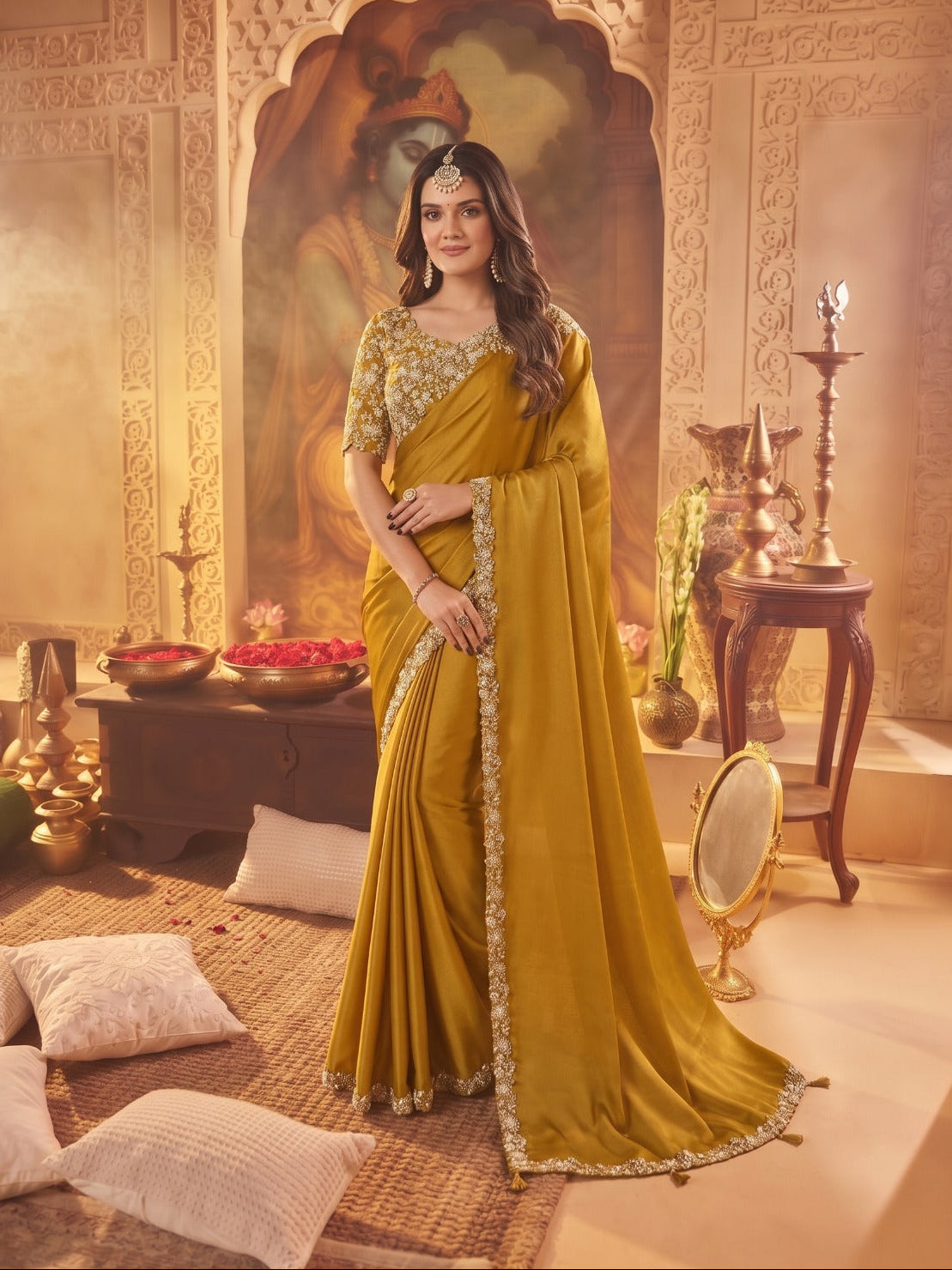 A woman models a mustard yellow saree with gold embroidery in a warm, traditional Indian room setting with decorative pillars, lamps, and cushions.