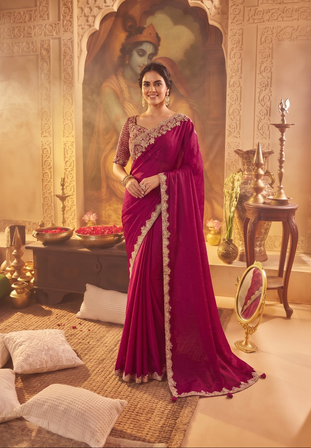 A woman models a deep magenta pink saree with gold embroidered borders and a matching embellished blouse in a warm, traditional Indian room setting.
