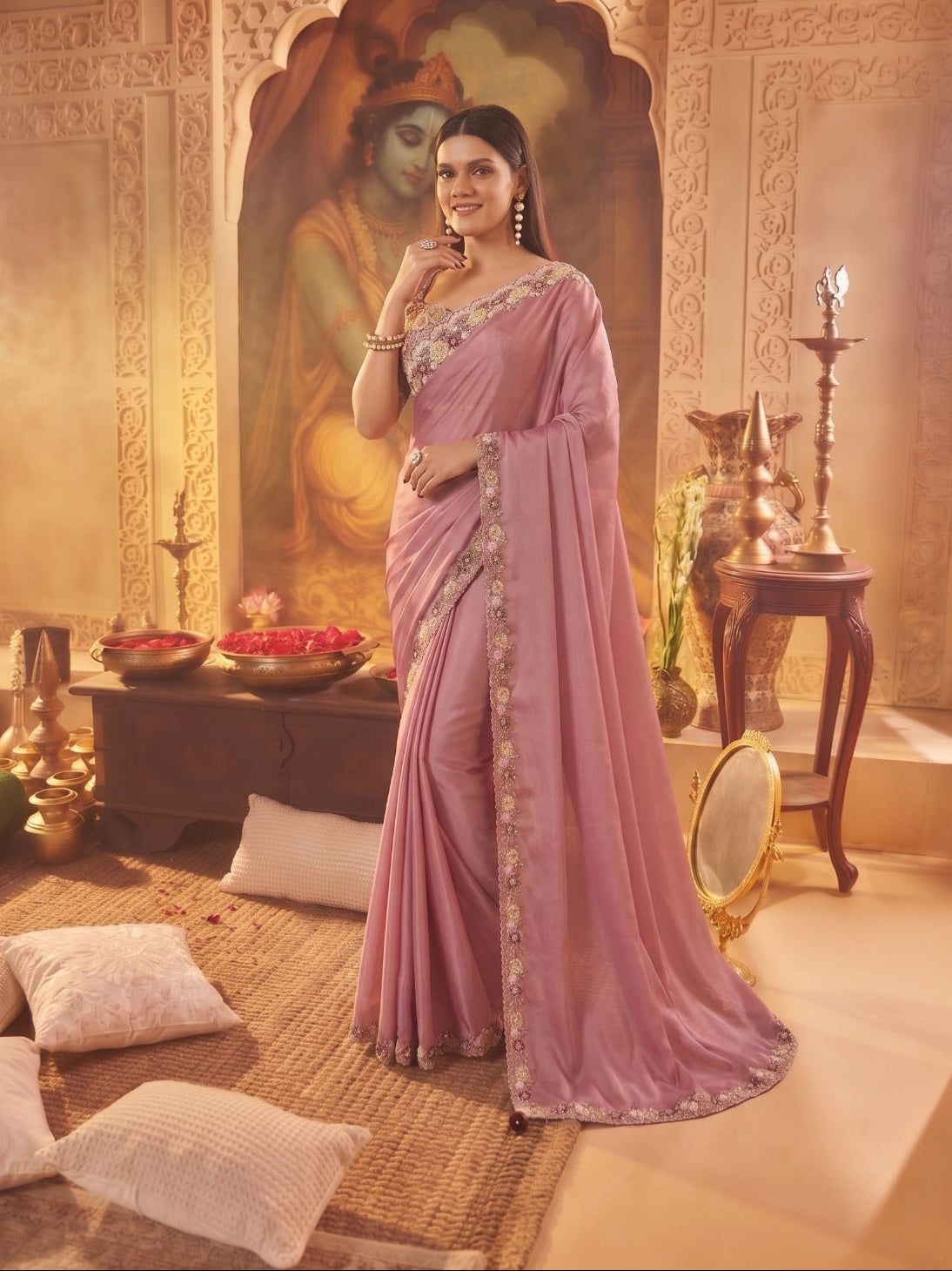 A woman models a lavender pink saree with gold embroidered borders in a traditional Indian room setting with warm lighting and decorative elements.