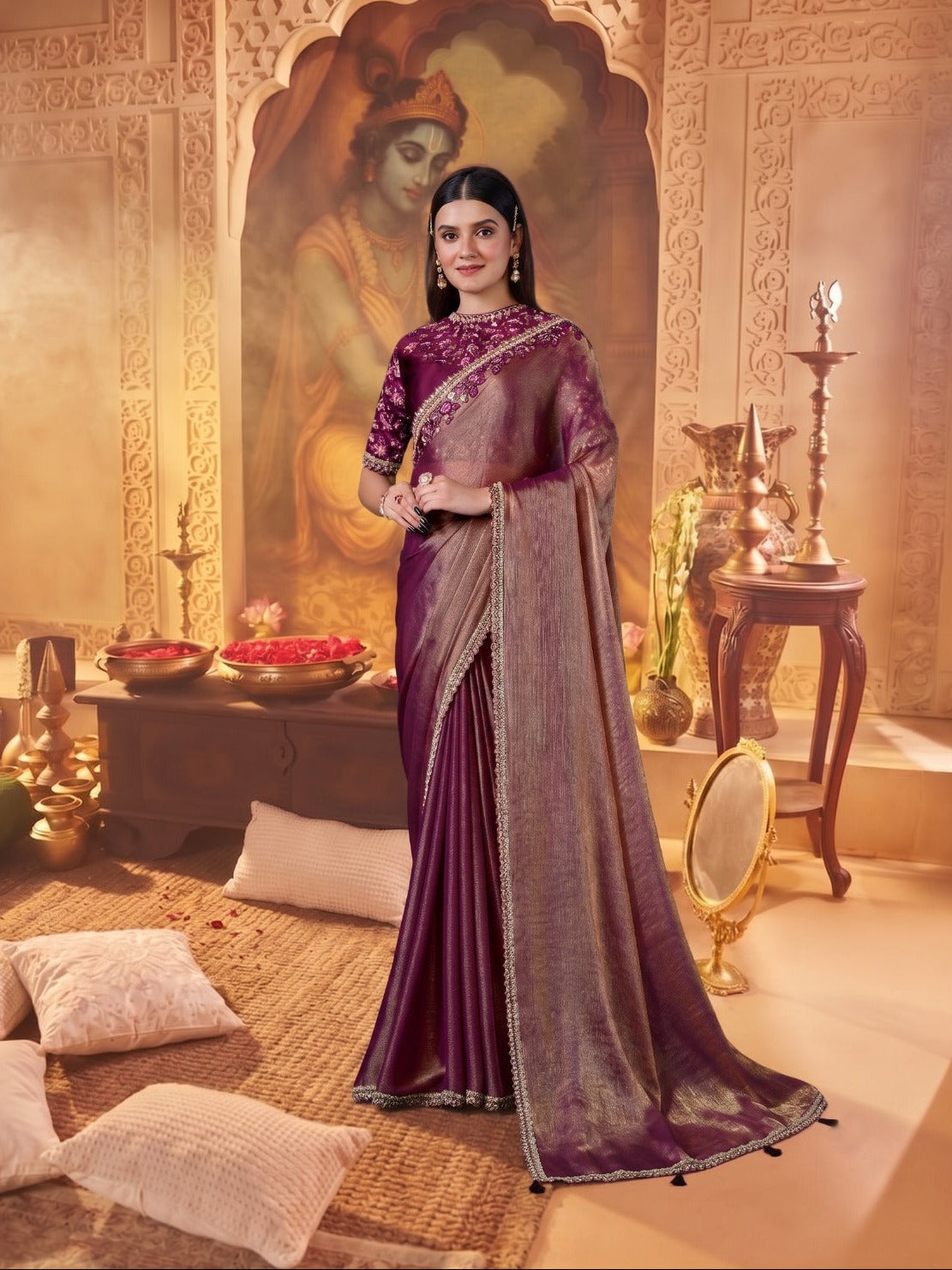 A woman models a purple and gold saree in an ornate, warm-toned Indian-style room with traditional decor, pillows, and a large framed painting in the background.