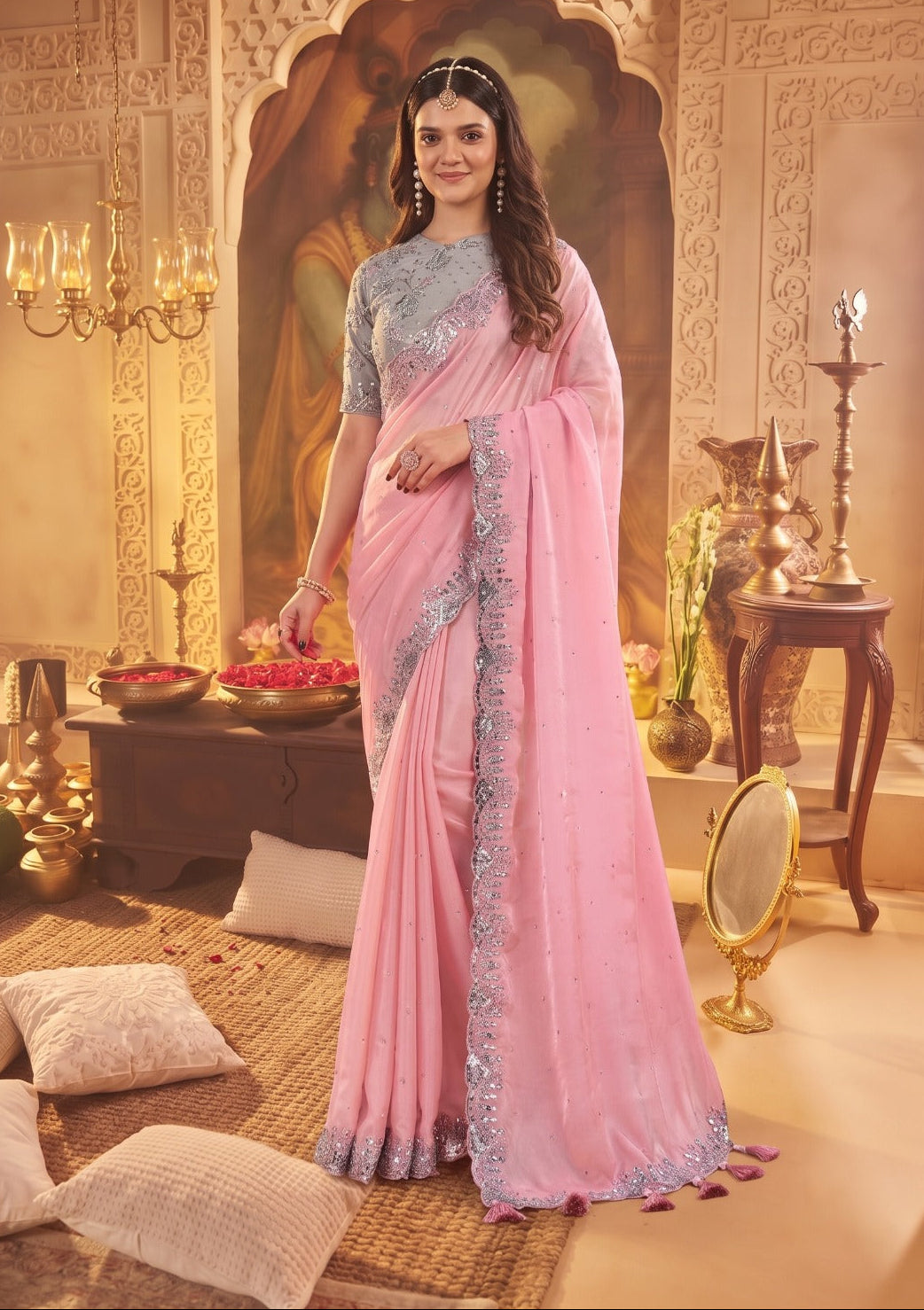 A woman models a light pink saree with a light blue embroidered blouse and border, standing in an ornate, warm-toned traditional room with decorative pillars, lamps, and cushions.