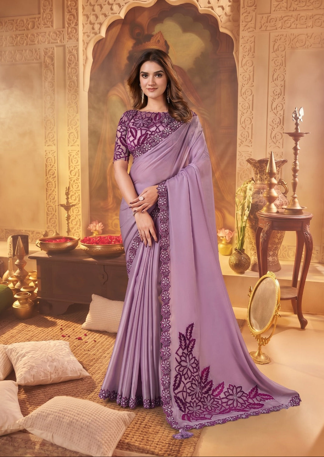 A woman models a lavender purple saree with a matching embroidered blouse in an ornate, warm-toned Indian-style room. The saree features a wide border with purple floral embroidery and a matching embroidered pallu.