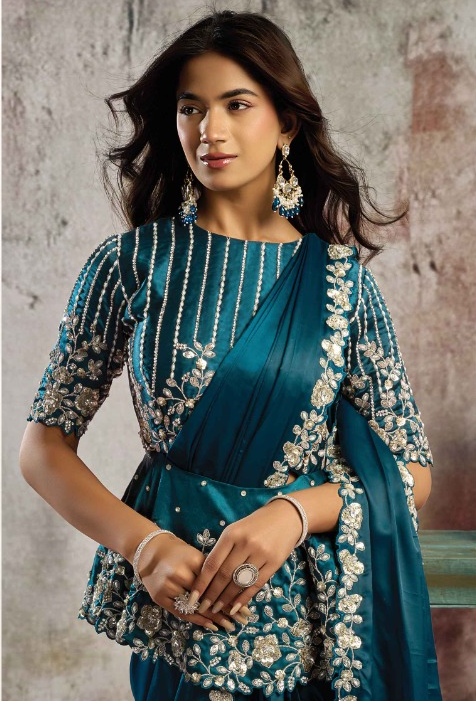 A woman modeling a teal blue traditional outfit with intricate silver and white floral embroidery, featuring a peplum-style skirt and matching draped fabric.