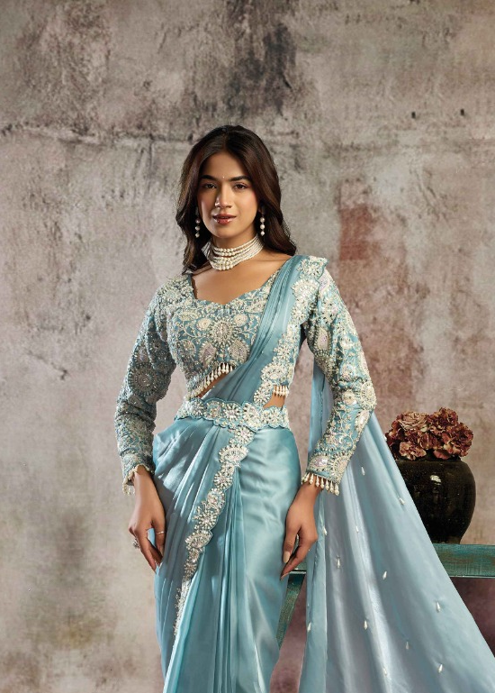 A woman modeling a light blue traditional Indian outfit with intricate silver embroidery, a matching draped dupatta, and a pearl necklace, posed against a textured gray wall.