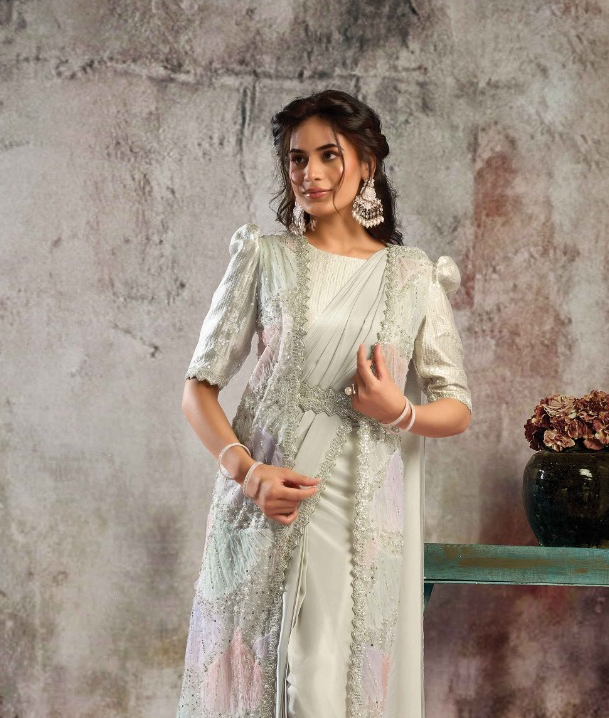 A woman models a traditional South Asian outfit consisting of a white satin saree and a matching heavily embroidered light silver-gray jacket, standing against a textured gray wall.