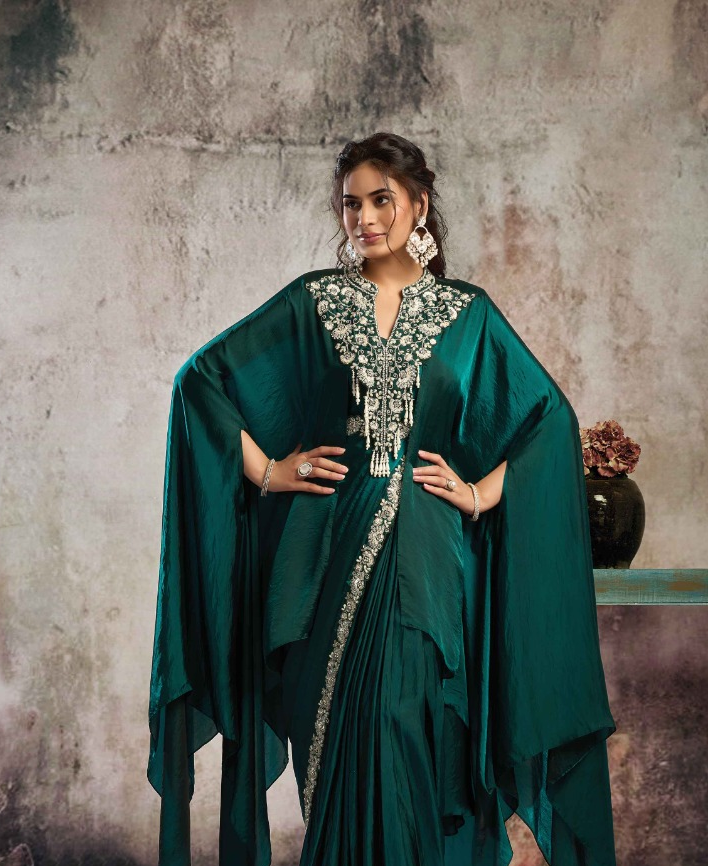 A woman models a dark teal green traditional Indian salwar suit with a draped cape-style jacket and heavy silver mirror work embroidery on the neckline and border, set against a textured gray studio backdrop.
