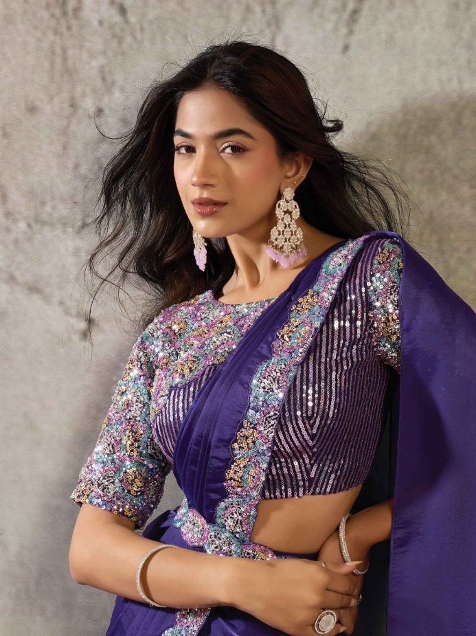A woman wearing a purple traditional outfit with heavily embellished floral embroidery and sequins on the blouse, posed against a textured gray wall.