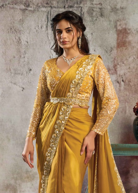 A woman modeling a mustard yellow saree with ornate gold embroidery on the blouse and border, posed against a textured gray wall.
