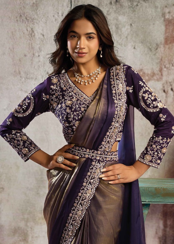 A woman modeling a traditional Indian outfit featuring a deep purple embroidered kurta with a matching metallic bronze saree, posed against a textured gray wall.