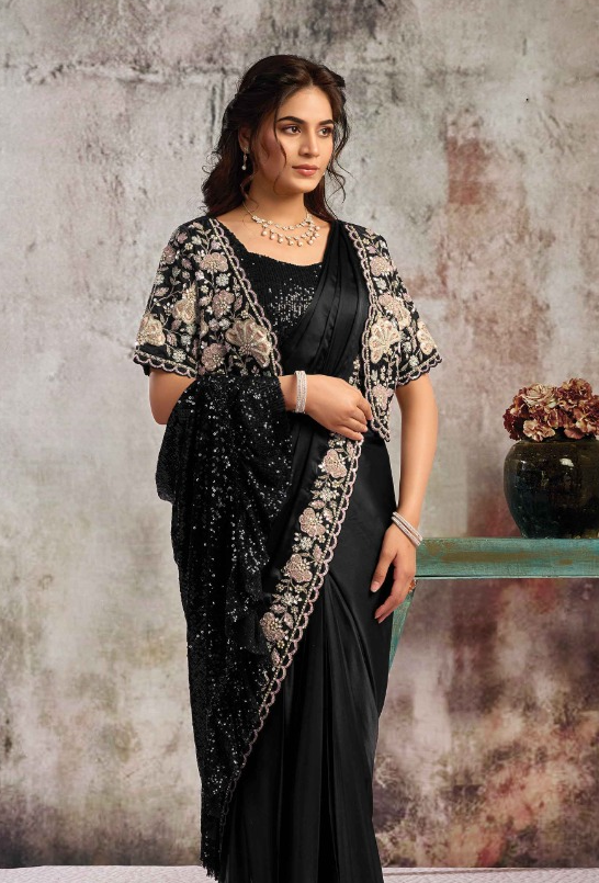 A woman modeling a black saree with intricate beige and gold floral embroidery on the border and sleeves, paired with a matching black embellished blouse, set against a textured gray studio backdrop.