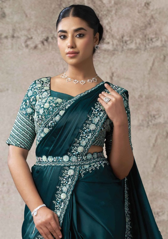 A woman modeling a dark teal green saree with intricate silver and white embroidery on the blouse and border, set against a textured beige wall.