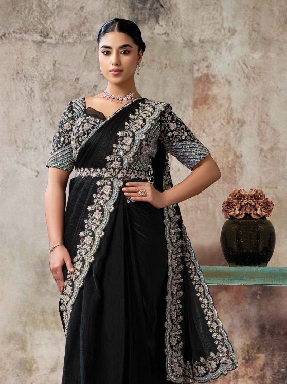 A woman models a black sari with intricate silver and white floral embroidery along the border and sleeves, set against a textured beige wall with a decorative vase in the background.