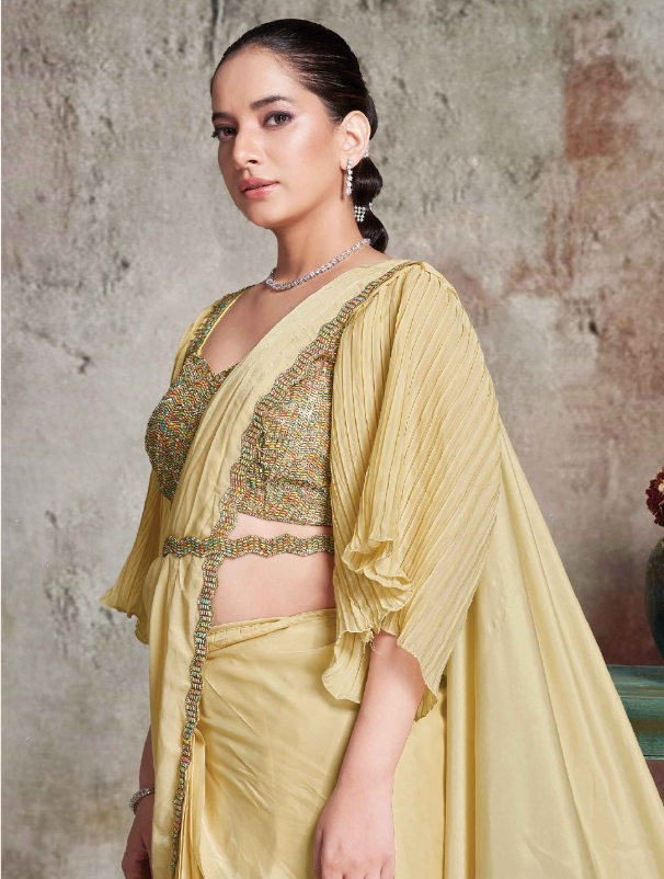 A woman modeling a light yellow georgette saree with heavily embroidered gold and green blouse, draped elegantly against a textured gray wall.