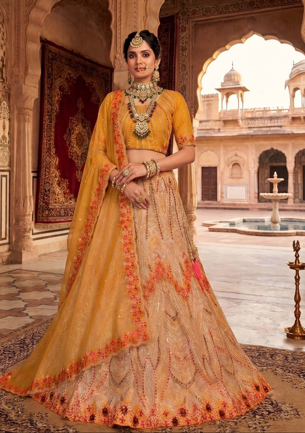A woman models a traditional Indian lehenga choli outfit in a grand palace courtyard. The outfit consists of a mustard yellow short-sleeve blouse with a heavily embroidered beige and orange skirt and dupatta.