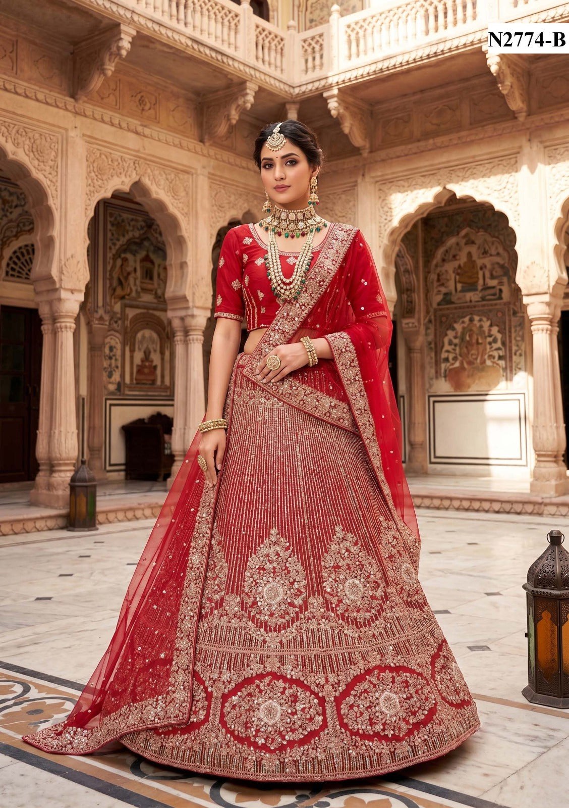 A woman stands in a carved stone courtyard wearing an ornate red and gold embroidered traditional outfit with a matching dupatta, heavy jewelry, and a decorative skirt with large circular motifs.