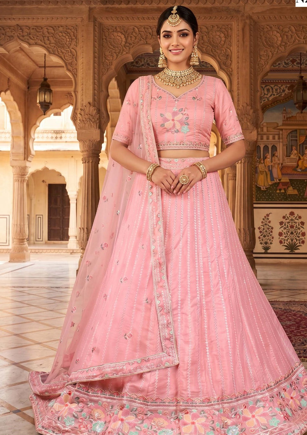 A woman stands in an ornate indoor courtyard wearing a pink traditional embroidered outfit with a flared skirt, matching blouse, and draped dupatta. The setting features carved arches and decorative walls, and the product is shown full length.