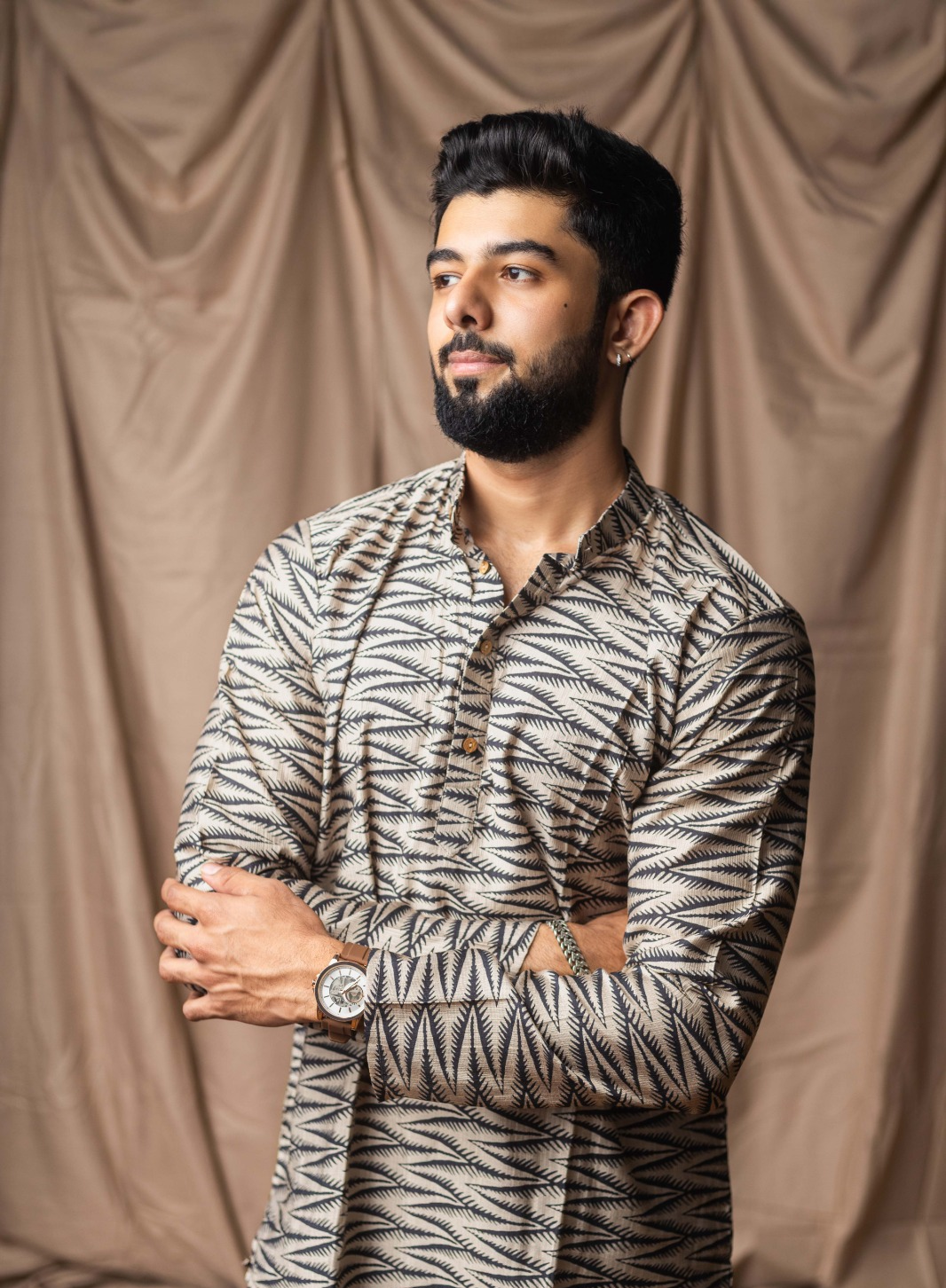 A man with a beard and styled hair wearing a long-sleeve kurta with a black and beige geometric pattern, standing against a draped brown fabric backdrop.
