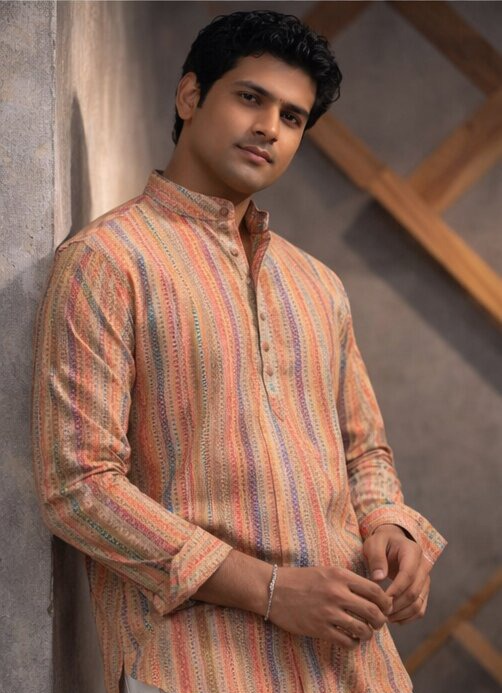 A man wearing a long-sleeve, mandarin-collar kurta with vertical stripes in orange, beige, and blue, leaning against a textured gray wall.