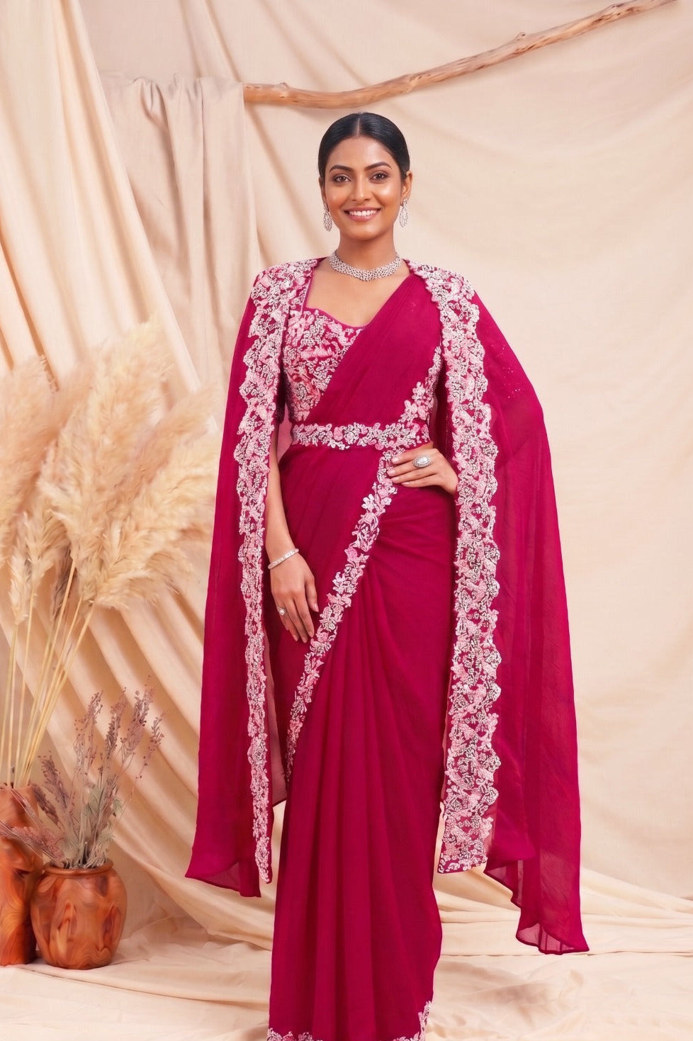 A woman models a deep magenta pink saree with intricate white floral embroidery along the border, draped over a matching embroidered sheer cape. The setting features a draped beige fabric backdrop with decorative pampas grass in wooden pots.
