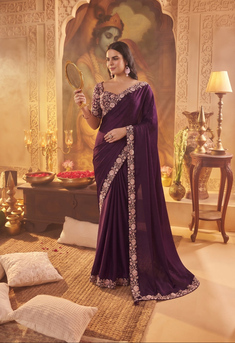 A woman models a deep purple saree with intricate silver floral embroidery along the border and blouse, standing in a richly decorated Indian-style room with warm golden lighting.
