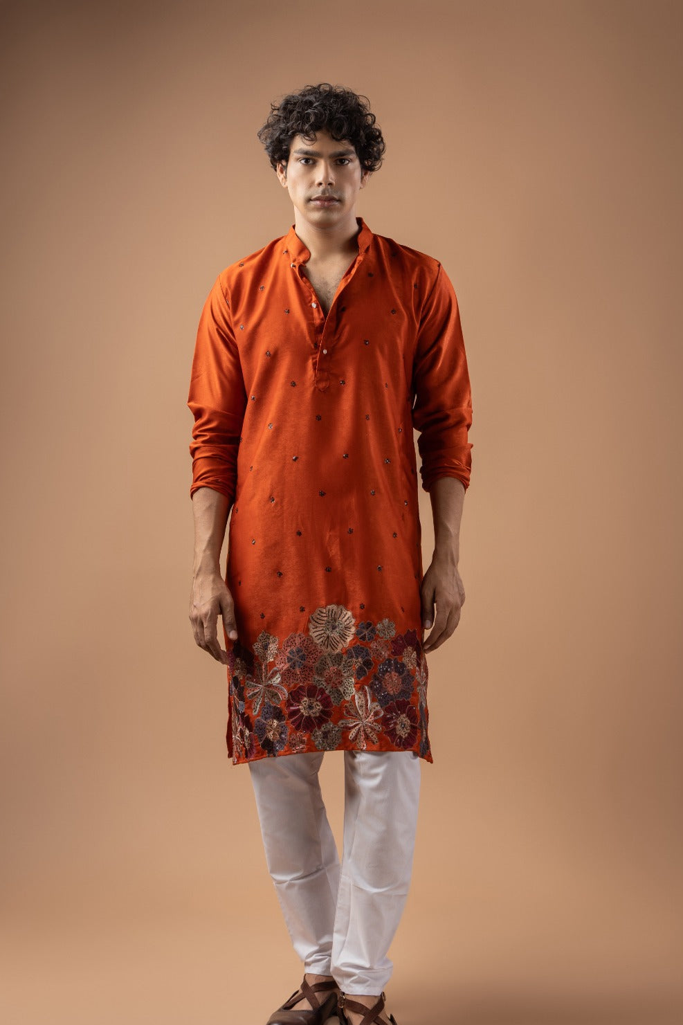 A man modeling a bright orange kurta with a floral embroidered hem, paired with white trousers and brown sandals, set against a solid warm brown studio background.