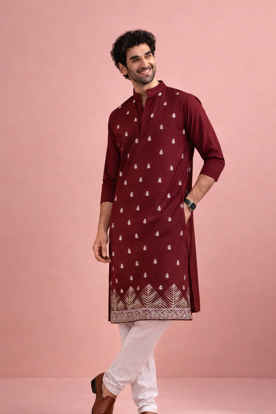 A man modeling a maroon long kurta with white printed motifs and a decorative border, paired with white pants and brown shoes, against a solid pink studio backdrop.