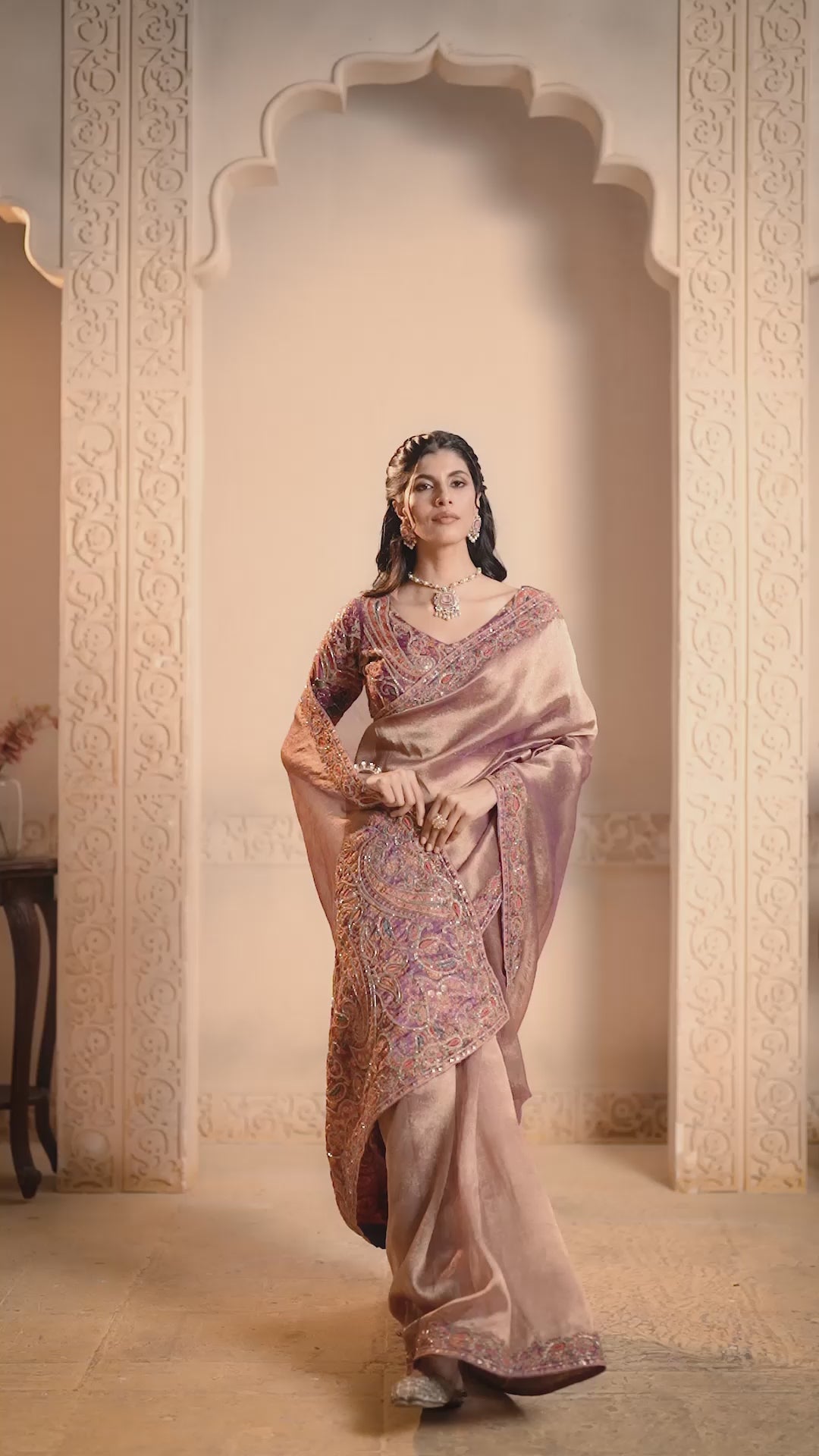 A woman models a purple silk saree with intricate gold, orange, and red paisley embroidery along the border and sleeves.